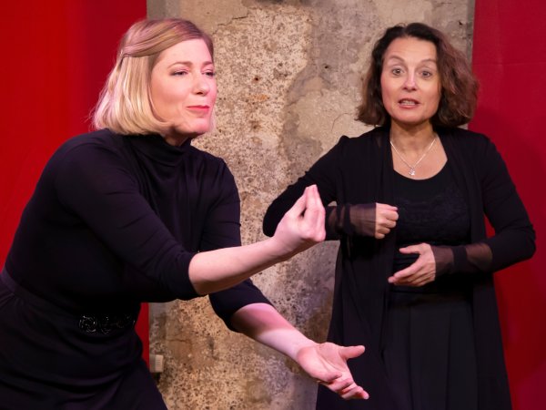 zwei Frauen auf Bühne schwarz gekleidet, gestikulieren