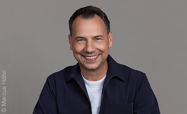 Foto Sebastian Fitzek: Lächelnde Person mit dunkler Jacke über weißem T-Shirt vor grauem Hintergrund.