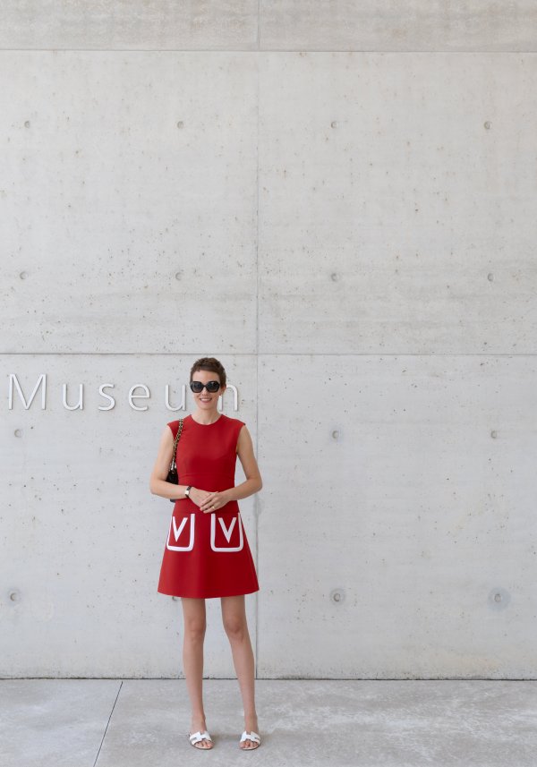 Person in rotem Kleid und Sonnenbrille steht lächelnd vor einer Betonwand mit der Aufschrift „Museum“.