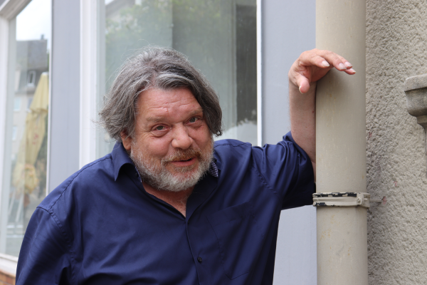 Person mit grauem Haar und Bart im dunkelblauen Hemd lehnt lächelnd an einem Fallrohr vor einer Hausfassade.