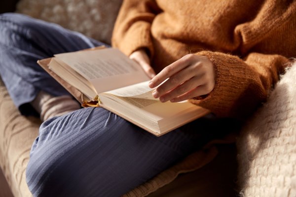 Person in Strickpullover sitzt im Schneidersitz auf einem Sofa und blättert in einem aufgeschlagenen Buch, dass auf ihrem Schoß liegt.