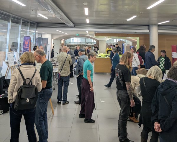 Viele Menschen stehen und warten in einer belebten Bibliothekshalle mit Infotafeln und Ausstellungstischen.