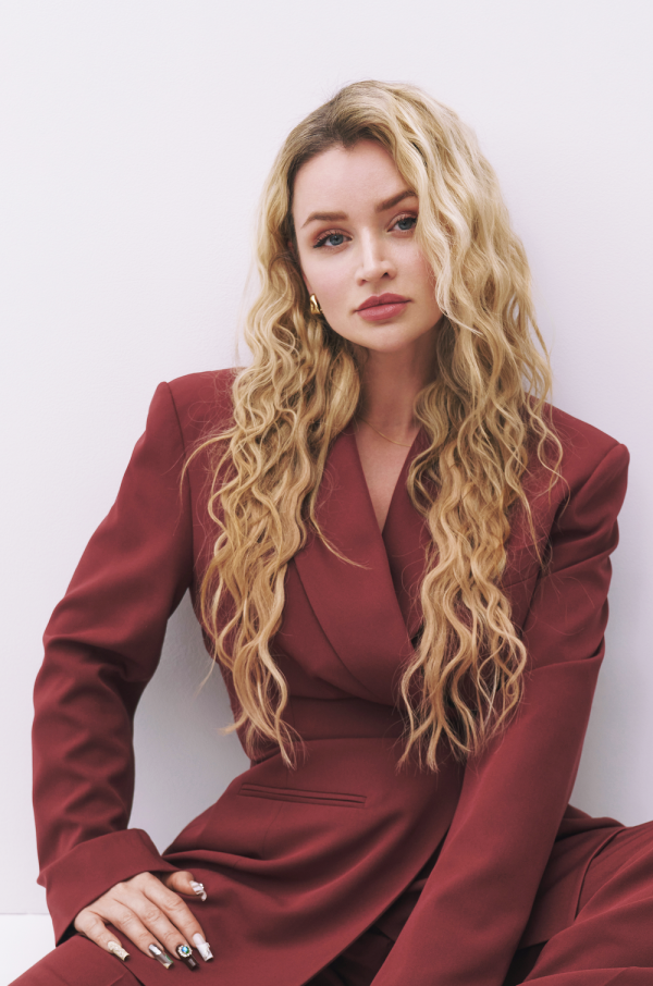 Person mit langen blonden Locken und dunkelrotem Blazer sitzt vor einer hellen Wand.