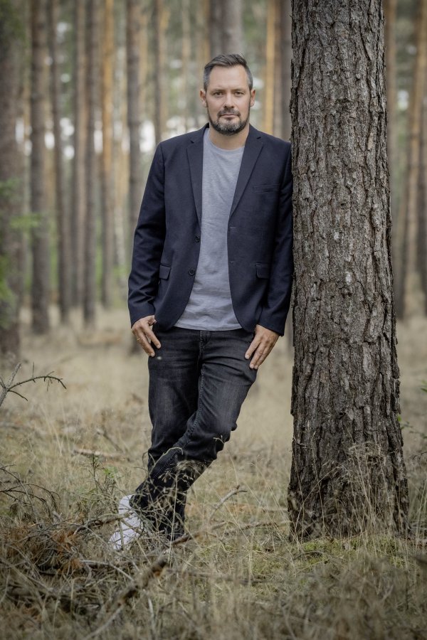 Person mit dunklem Sakko und grauem T-Shirt lehnt an einem Baumstamm in einem Kiefernwald.