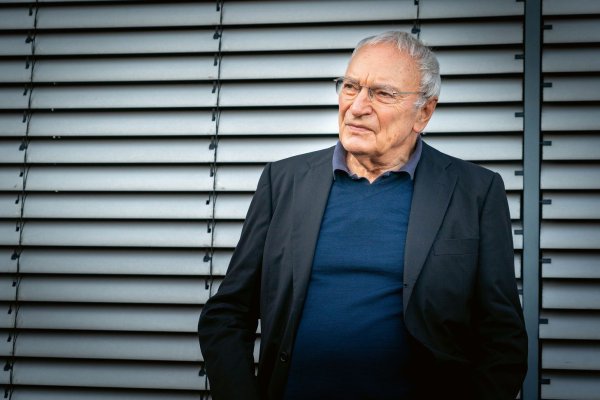 Uwe Timm in dunklem Sakko und blauem Pullover steht vor einer Lamellenwand.