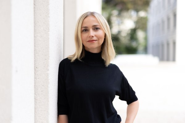 Porträt einer Frau mit blondem Haar und schwarzem Rollkragenpullover, die an einer hellen Fassade steht und direkt in die Kamera blickt.