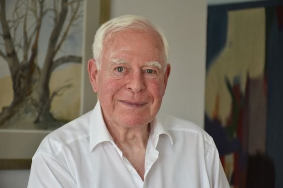 Ein älterer Mann (Ernst Krakenberger) mit weißen Haaren und weißem Hemd lächelt vor einer Wand mit gerahmten Gemälden.