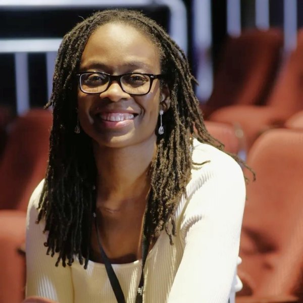 Sharon Dodua Otoo: Lächelnde Person mit Brille und Dreadlocks sitzt zwischen roten Sitzreihen in einem Theater- oder Kinosaal.