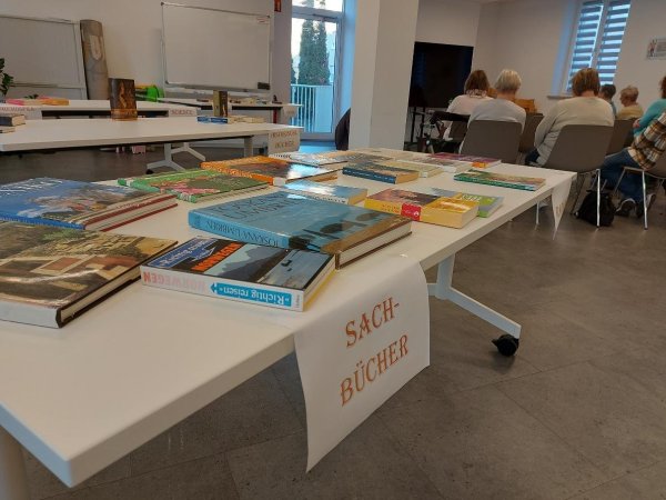 Tische mit sortierten Büchern, unter anderem mit dem Schild „Sachbücher“, im Hintergrund sitzen Personen im Raum.
