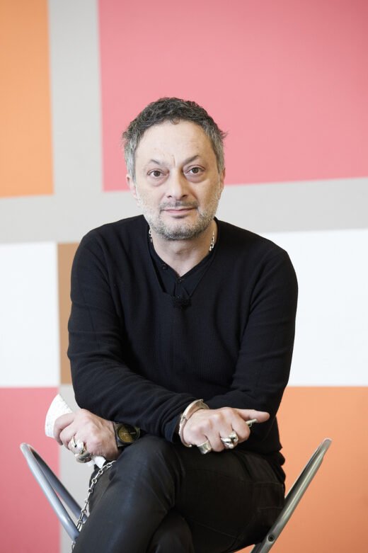 Ein Mann mit dunklem Pullover und mehreren Ringen sitzt vor einer bunten, geometrisch gemusterten Wand.