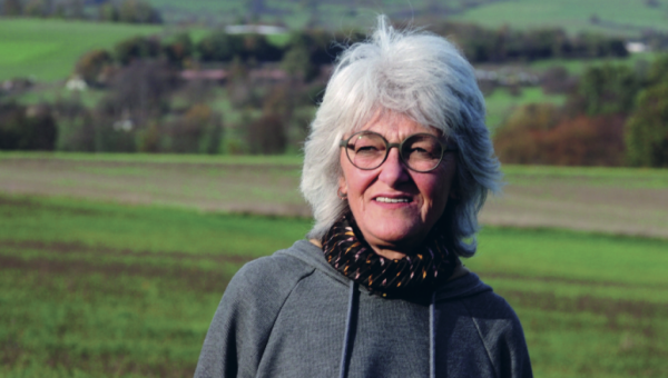 Person mit grauen Haaren und Brille steht lächelnd vor einer grünen Hügellandschaft.