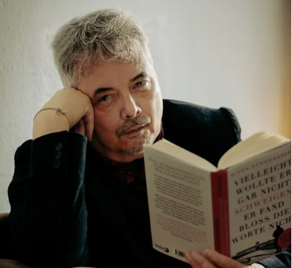 Eine Person mit grauen Haaren stützt den Kopf auf die Hand und hält ein aufgeschlagenes Buch in der anderen Hand.