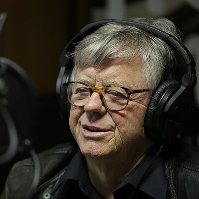 Fitzgerald Kusz: Ältere Person mit Brille und Kopfhörern spricht in ein Mikrofon in einem Tonstudio.