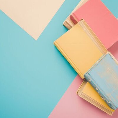 Mehrere Bücher mit pastellfarbenen Einbänden in Rosa, Gelb und Blau liegen gestapelt auf einem geometrisch gemusterten Hintergrund in Türkis, Creme und Rosa.