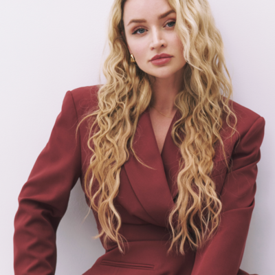 Person mit langen blonden Locken und dunkelrotem Blazer sitzt vor einer hellen Wand.