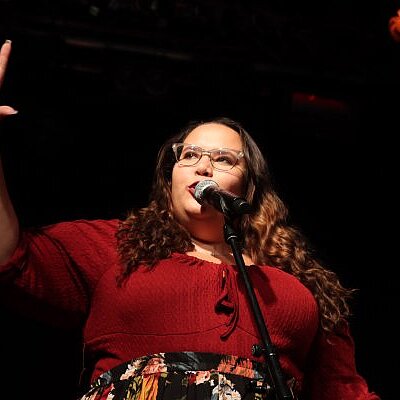 Auf dem Bild ist eine Frau in einem roten Oberteil an einem Mikrofon zu sehen, die einen Arm erhoben hat und den Horizont zeigt.