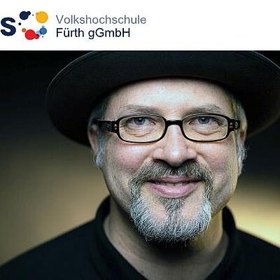 Porträt von Benjamin Krauss mit Hut, Brille und grauem Bart, darüber das Logo der Volkshochschule Fürth gGmbH.