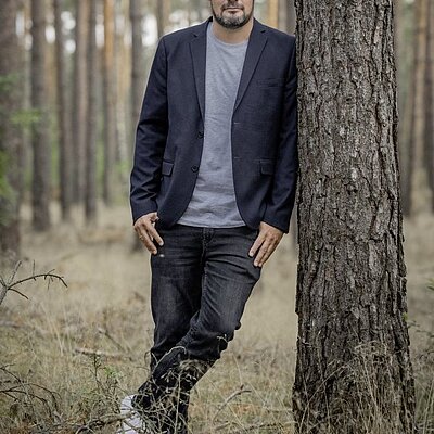 Person mit dunklem Sakko und grauem T-Shirt lehnt an einem Baumstamm in einem Kiefernwald.
