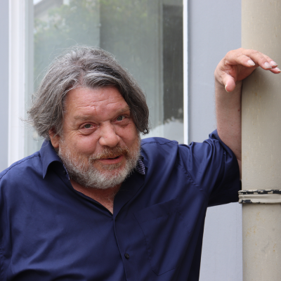Person mit grauem Haar und Bart im dunkelblauen Hemd lehnt lächelnd an einem Fallrohr vor einer Hausfassade.