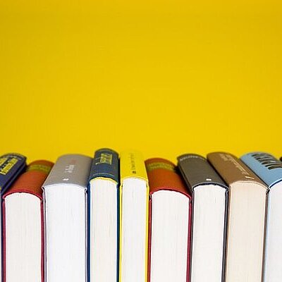Eine Reihe unterschiedlich farbiger Buchrücken steht aufrecht vor einem gelben Hintergrund.