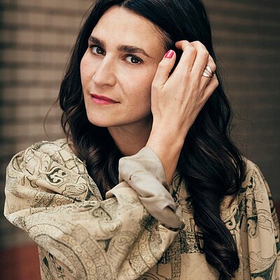 Portrait Yade Önder: Person mit dunklem, welligem Haar posiert nachdenklich mit der Hand am Kopf vor einer Backsteinwand.