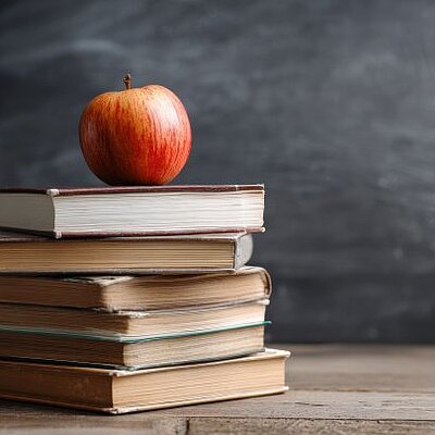 Ein roter Apfel liegt auf einem Stapel gebundener Bücher vor einer Tafel im Hintergrund.