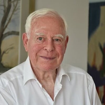 Ein älterer Mann (Ernst Krakenberger) mit weißen Haaren und weißem Hemd lächelt vor einer Wand mit gerahmten Gemälden.
