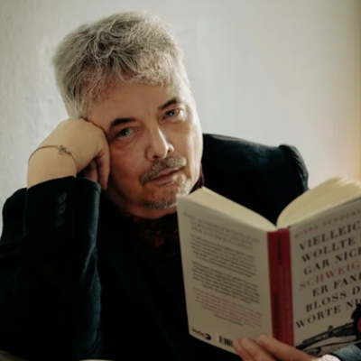 Eine Person mit grauen Haaren stützt den Kopf auf die Hand und hält ein aufgeschlagenes Buch in der anderen Hand.