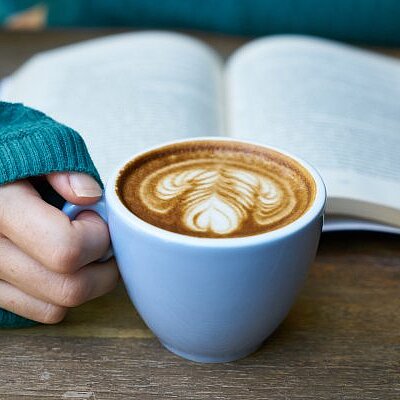 Eine Hand hält eine Tasse Kaffee mit Latte-Art vor einem aufgeschlagenen Buch auf einem Holztisch.