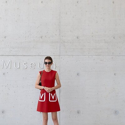 Person in rotem Kleid und Sonnenbrille steht lächelnd vor einer Betonwand mit der Aufschrift „Museum“.
