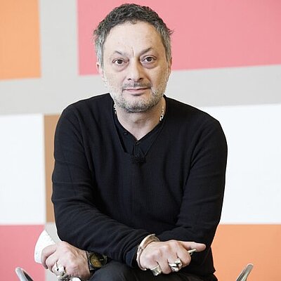 Ein Mann mit dunklem Pullover und mehreren Ringen sitzt vor einer bunten, geometrisch gemusterten Wand.