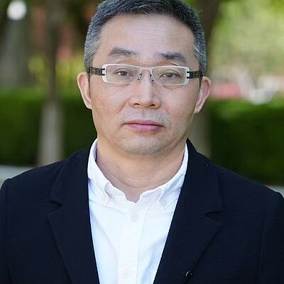 Der Schriftsteller Dong Xi: Porträt einer Person mit Brille, weißem Hemd und dunklem Sakko vor grünem Hintergrund im Freien.