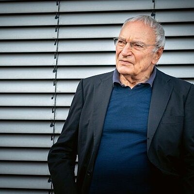 Uwe Timm in dunklem Sakko und blauem Pullover steht vor einer Lamellenwand.