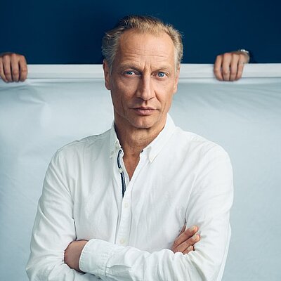 Portrait des Autors Victor Schefé. Er steht in weißem Hemd vor einem blau, weißen Hintergrund.