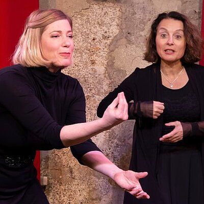 zwei Frauen auf Bühne schwarz gekleidet, gestikulieren