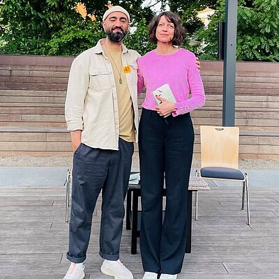 Portrait von Louise Brown (rechts) und Cihan Morsünbül (links). Frau Brown trägt ein pinkes Oberteil und eine dunkle Hose. Herr Morsünbül trägt eine helle Jacke und Kappe.