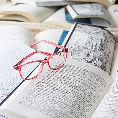Eine rosa Brille liegt auf einem aufgeschlagenen Buch inmitten mehrerer geöffneter Bücher mit bedruckten Seiten.