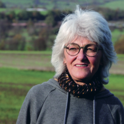 Person mit grauen Haaren und Brille steht lächelnd vor einer grünen Hügellandschaft.