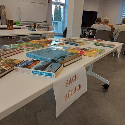 Tische mit sortierten Büchern, unter anderem mit dem Schild „Sachbücher“, im Hintergrund sitzen Personen im Raum.