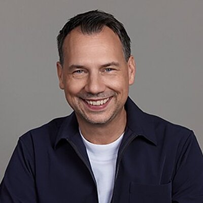 Foto Sebastian Fitzek: Lächelnde Person mit dunkler Jacke über weißem T-Shirt vor grauem Hintergrund.