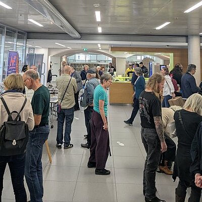 Viele Menschen stehen und warten in einer belebten Bibliothekshalle mit Infotafeln und Ausstellungstischen.
